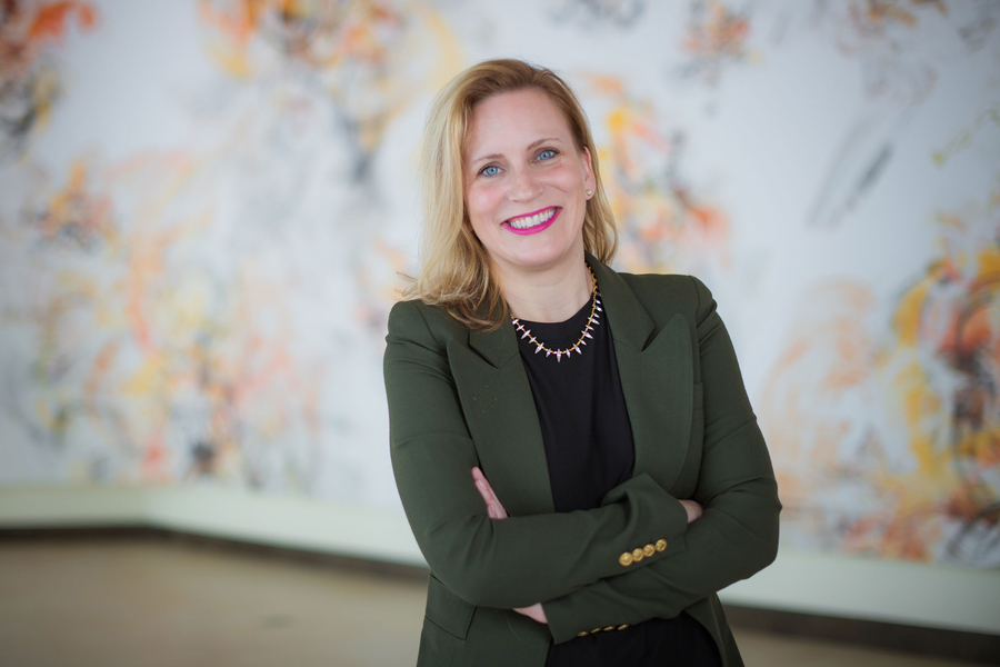 A smiling woman in a green blazer with arms crossed stands in front of a large abstract painting in warm orange and gold tones