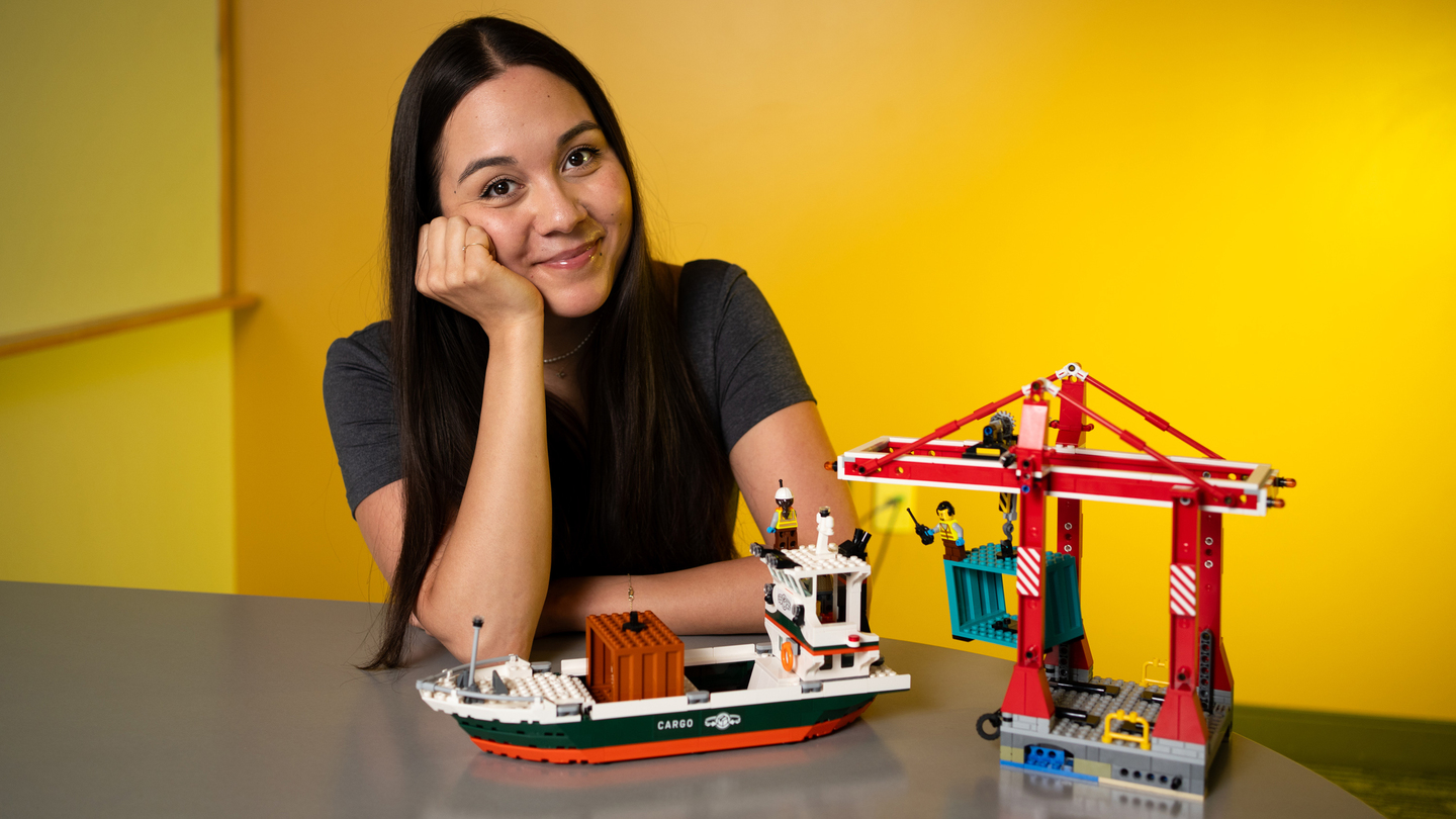 A smiling woman sitting at a desk beside a Lego model of a cargo ship and a port crane against a solid yellow wall.