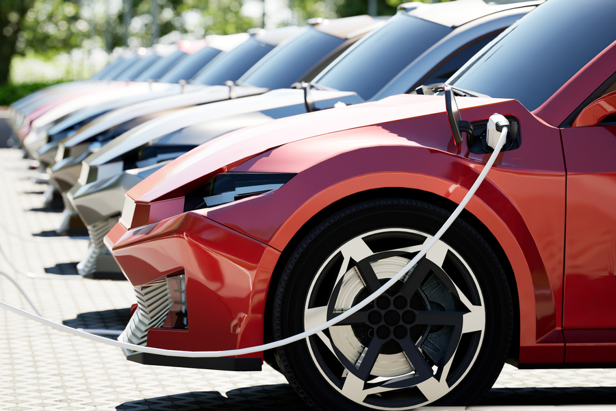 A row of electric vehicles plugged into charging cables in an outdoor lot, with a red EV in the foreground and additional cars receding into the background.