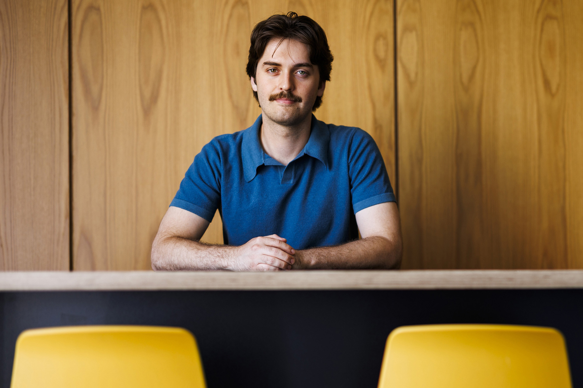Man seated behind a counter, wearing a blue polo shirt, with two yellow chairs in the foreground.