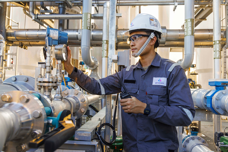 A Gradiant-branded technician in a hard hat and safety glasses inspects a pressure gauge on an industrial pipe system.