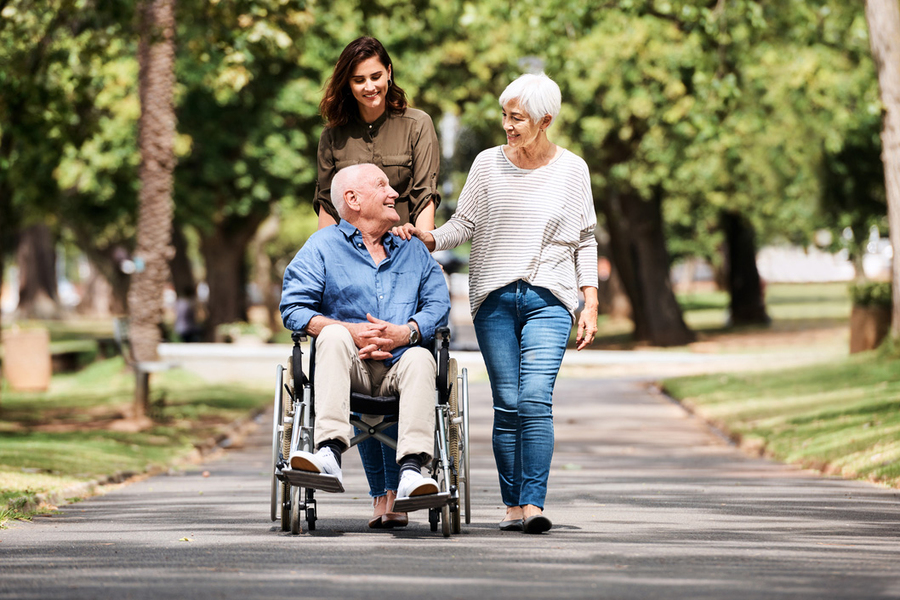 Two caregivers and an older man in a wheelchair sharing a cheerful moment while walking along a tree-lined path outdoors.