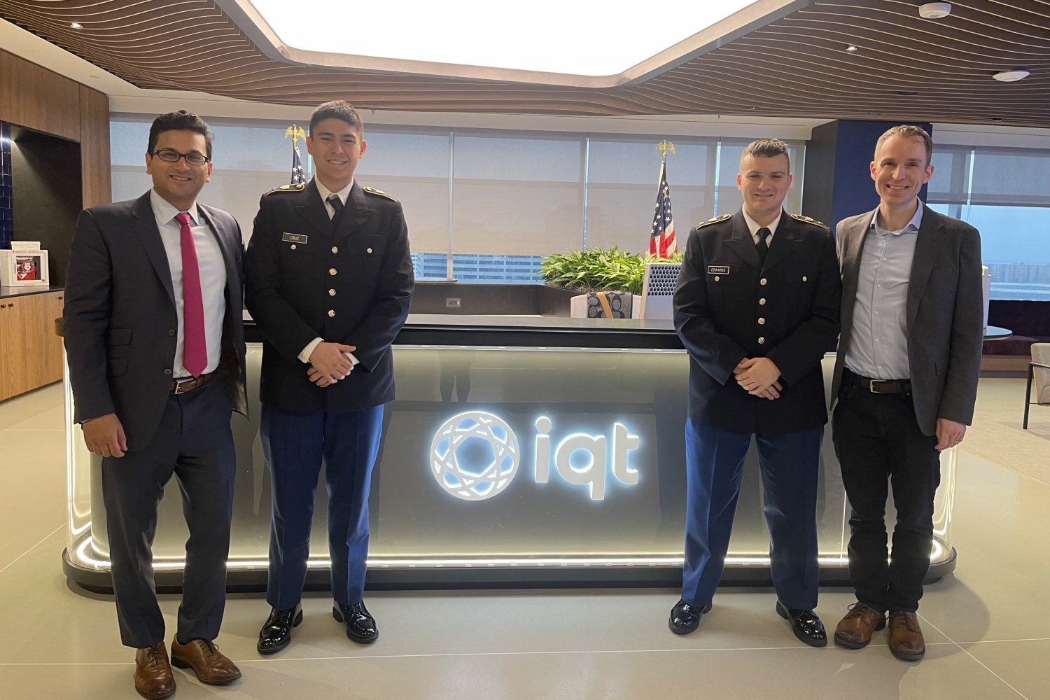 Five men stand before illuminated IQT logo in modern office: two in military uniforms, three in business suits, with cityscape visible through windows.