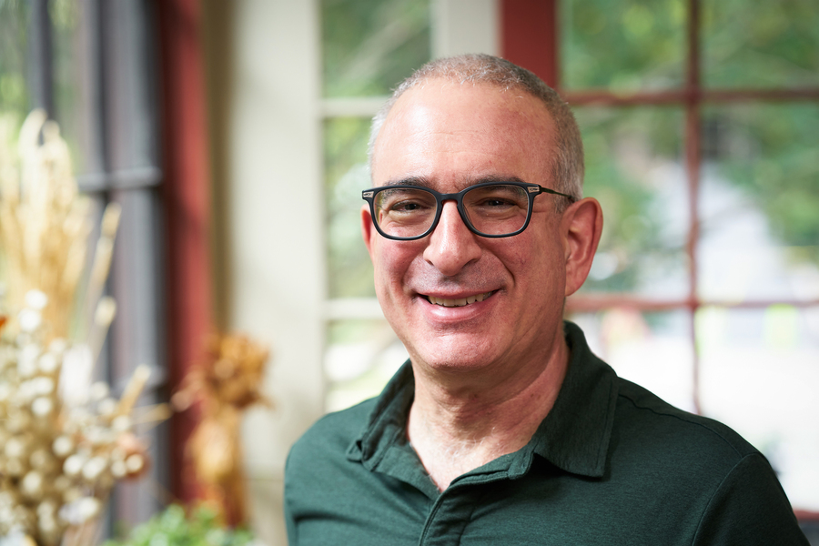 Person smiling and standing indoors near a window, with plants and soft natural light in the background.