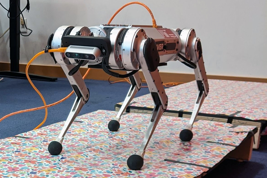 Four-legged robotic dog with white and reddish-brown body segments, black feet, and mounted sensors, tethered with orange cable on patterned floor indoors.