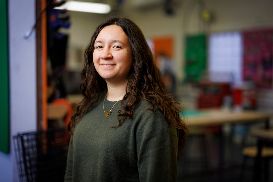 Woman with long dark wavy hair wearing olive green sweater, smiling at camera in classroom with colorful storage containers blurred in background.