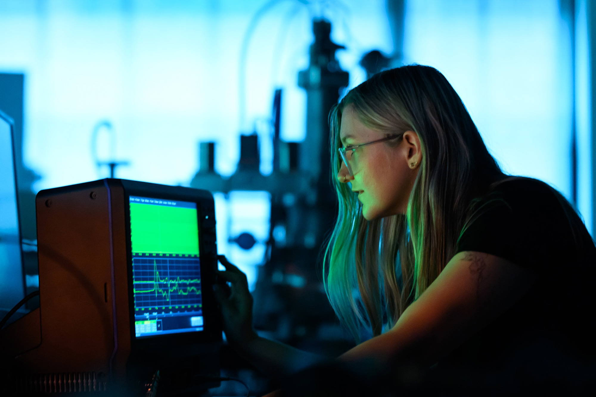 Researcher monitoring a waveform display on scientific equipment in a dimly lit laboratory.