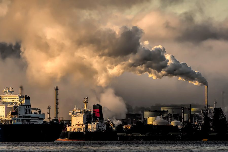 Industrial facility with large smoke plumes rising from chimneys near docked ships.