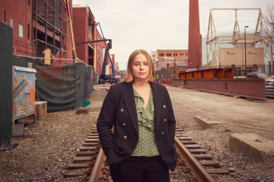 Portrait of a woman standing on railroad tracks amid an urban industrial construction site, wearing a black blazer and green blouse.