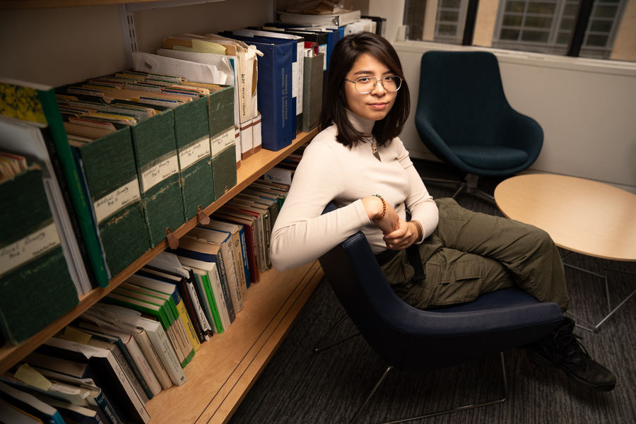 A woman in glasses and a cream turtleneck sits beside a floor-to-ceiling shelf of academic binders and research volumes in an office setting.