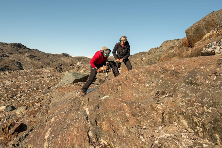 Two fieldwork researchers in protective gear using a rock drill to extract samples from ancient exposed bedrock in a remote, rocky landscape under a clear blue sky.