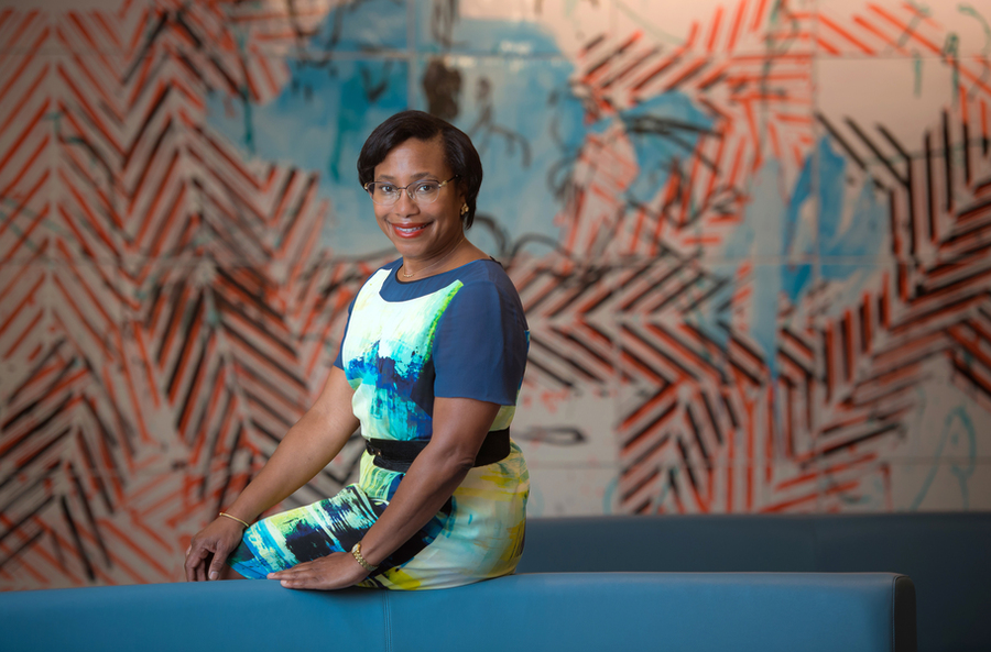 A woman in a colorful patterned dress sits on a blue bench in front of a bold abstract mural with orange and black zigzag patterns.