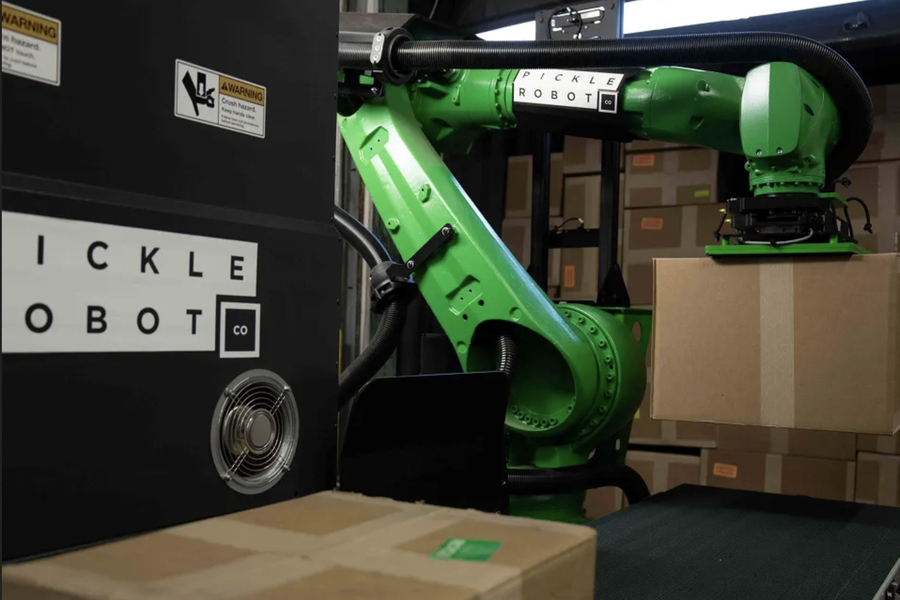 A green robotic arm lifting a cardboard box inside a workspace stacked with packages.