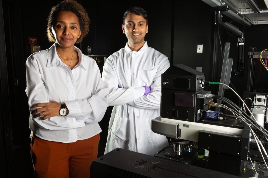 Two researchers in lab coats standing beside advanced microscopy equipment inside a dark laboratory.