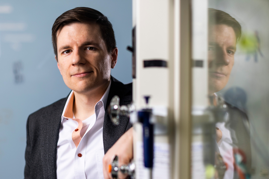 Professional portrait of man in dark blazer and white shirt standing before office glass with reflection visible.