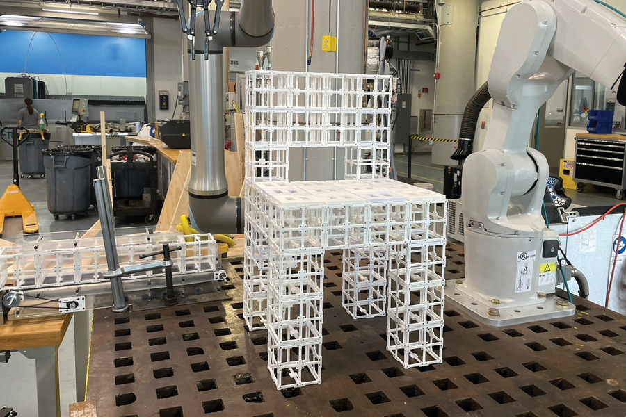 A grid‑like modular chair structure sits on a worktable in a robotics lab with robotic arms and tools around it.