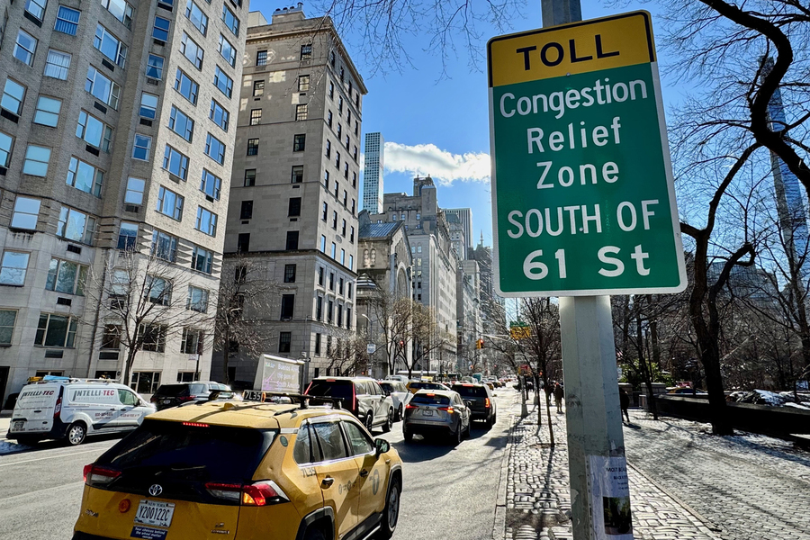 Yellow taxi and traffic on Manhattan street with green "Toll Congestion Relief Zone" sign and tall buildings under blue sky.