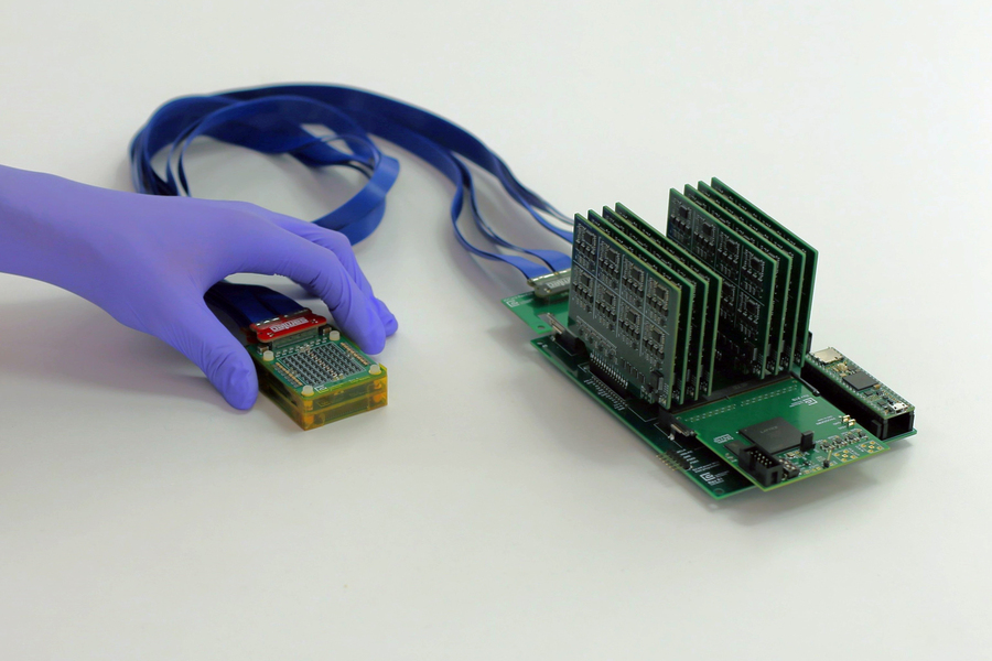 A gloved hand holding a small electronic module connected by ribbon cables to a larger circuit board.