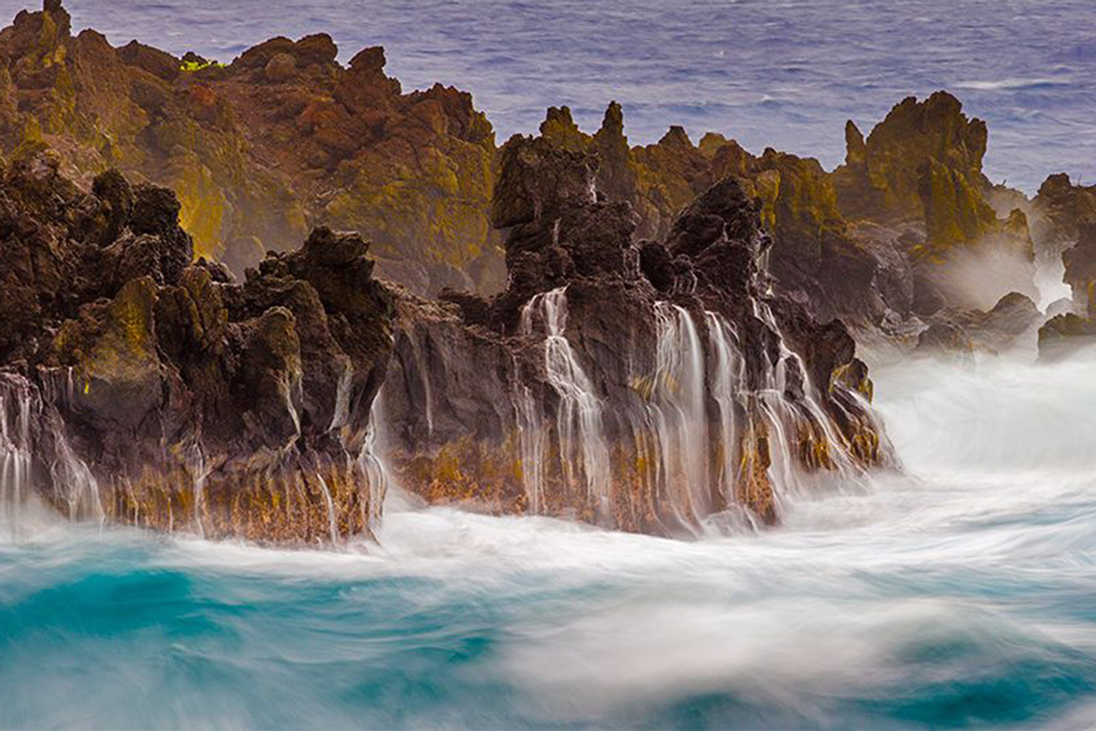 Long-exposure photograph of powerful ocean waves crashing against jagged volcanic lava rock formations on a Hawaiian coastline, with silky white surf and blue water.