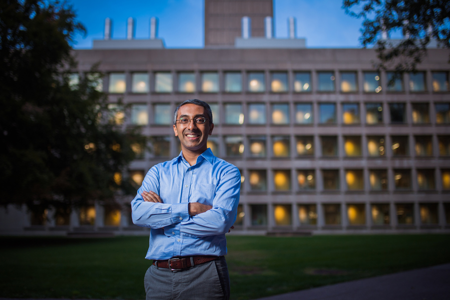 A smiling researcher in a blue button-down shirt stands with arms crossed on a campus lawn at dusk, with a large illuminated brutalist academic building in the background.