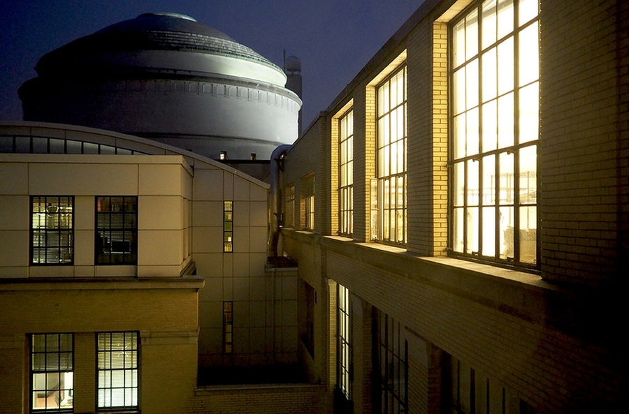 Night view of MIT's Great Dome rising above illuminated brick and industrial-style campus buildings with large factory-style windows