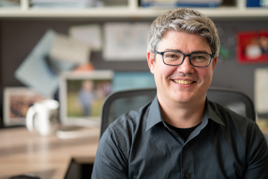 An MIT astrophysicist smiling at his desk, wearing glasses and a dark shirt, with papers and photos pinned to a bulletin board behind him.