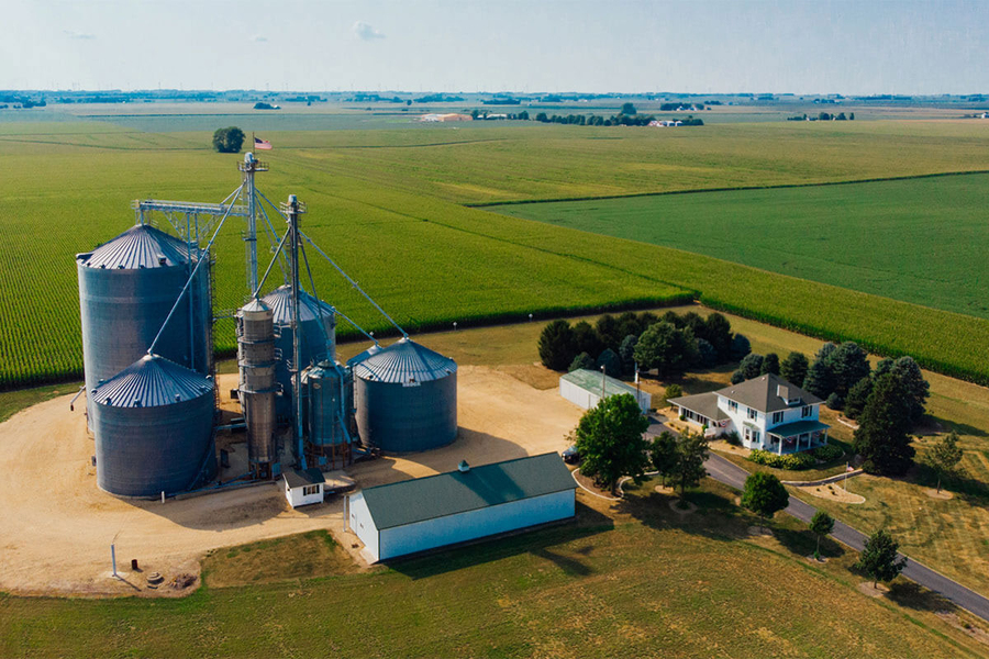 Aerial view of a midwestern farm with several large grain silos, a white farmhouse, and outbuildings surrounded by expansive green crop fields.