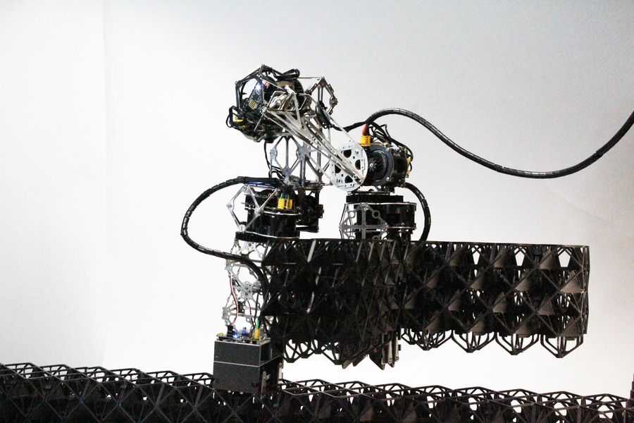 A black robotic arm assembling a structure made of repeating dark lattice pieces.