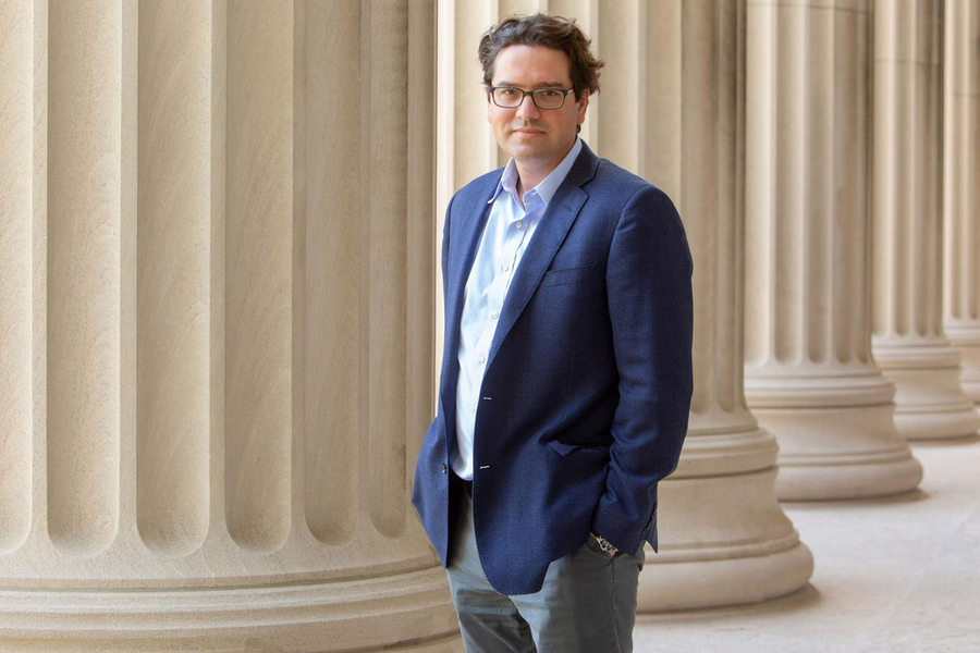 A man in a blue blazer and glasses stands with hands in pockets beside a row of tall stone columns in a classical campus corridor.