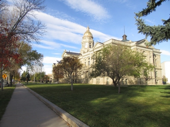 Exterior view of the Wyoming State Capitol building in Cheyenne on a sunny autumn day, with its gold dome visible above sandstone walls and surrounded by green lawns and fall-colored trees.