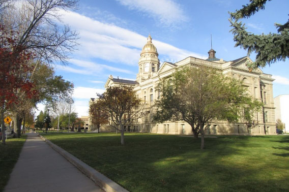 Exterior view of the Wyoming State Capitol building in Cheyenne on a sunny autumn day, with its gold dome visible above sandstone walls and surrounded by green lawns and fall-colored trees.