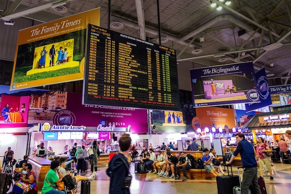 A bustling train station with people seated and walking. Overhead, a large digital display shows train schedules. Advertisements and eateries are visible, creating a lively atmosphere.