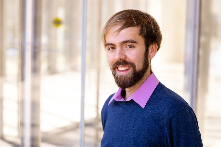 MIT researcher Matt Kearney smiles outdoors in front of a glass building exterior, wearing a navy sweater over a lavender collared shirt.