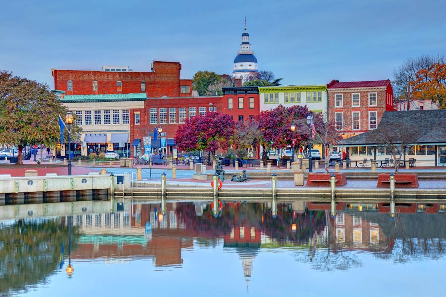Annapolis, Maryland harbor waterfront with colorful colonial storefronts reflected in calm water, and the Maryland State House dome rising above the hilltop.