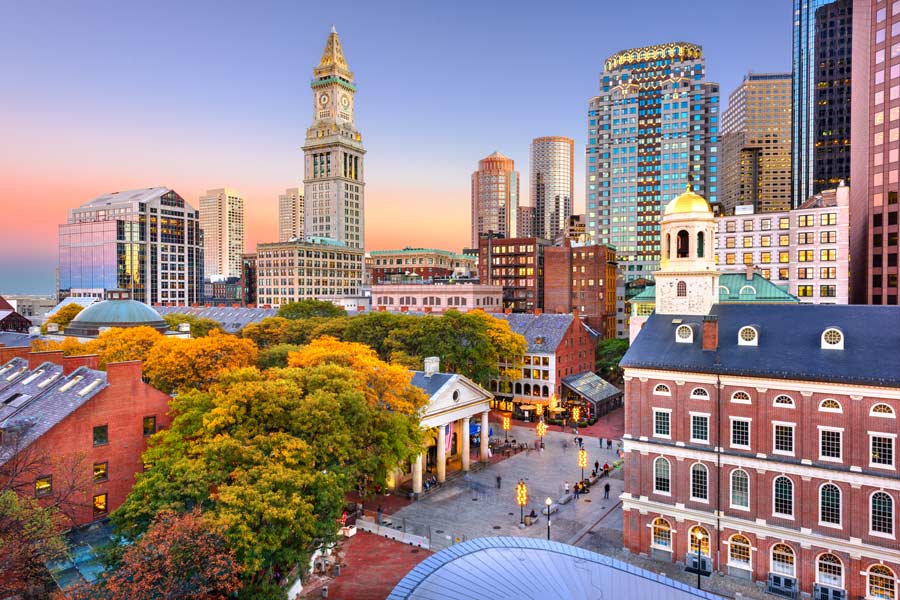 Boston, Massachusetts skyline at dusk featuring Faneuil Hall, the Custom House clock tower, and downtown skyscrapers amid autumn foliage.