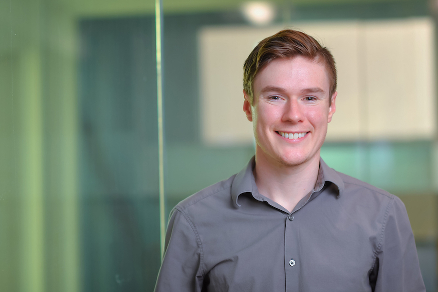 Headshot of a smiling young man in a gray button-down shirt against a soft green glass-panel background