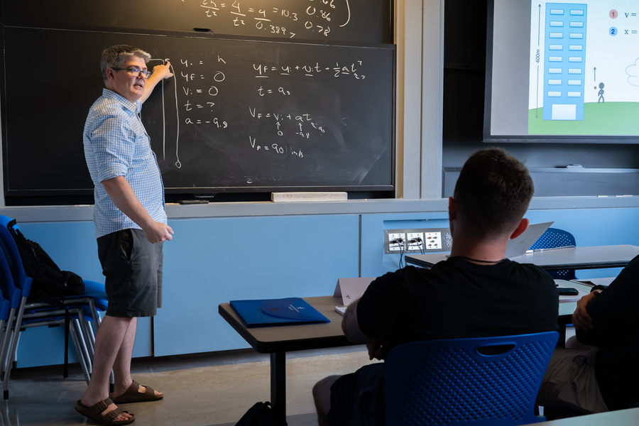 An instructor pointing to physics equations on a chalkboard while students sit at desks watching.