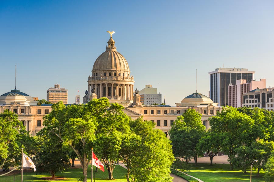 The Mississippi State Capitol building in Jackson, with its ornate stone dome topped by a golden eagle, rising above lush summer trees and green lawns.
