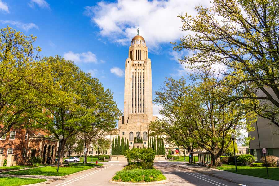 The Nebraska State Capitol building in Lincoln, framed by spring trees, with its distinctive Art Deco tower and bronze dome topped by a sower statue.