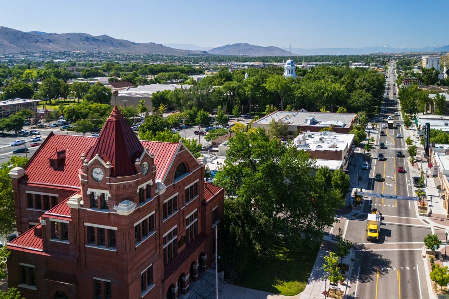 Downtown Carson City, Nevada with a Victorian-era red brick clock tower building in the foreground and the Nevada State Capitol dome visible down a tree-lined boulevard, with the Sierra Nevada mountains in the distance.