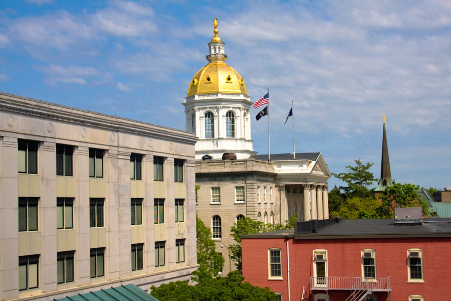 The New Hampshire State House in Concord, with its striking gold dome and white cupola flying the American flag, flanked by trees and neighboring buildings.