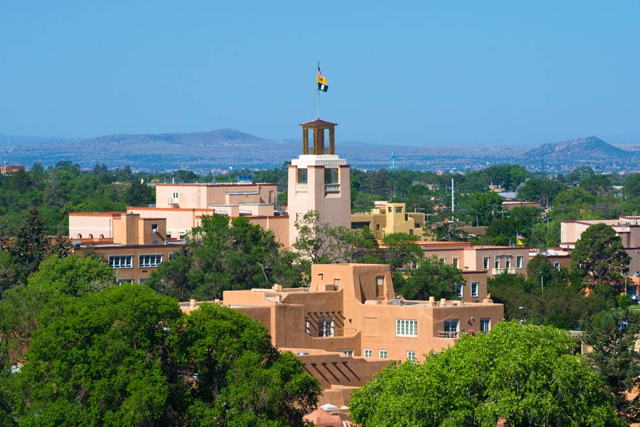 Downtown Santa Fe, New Mexico rooftops featuring traditional adobe Pueblo Revival architecture and a flagged tower, with the Jemez Mountains in the distance.