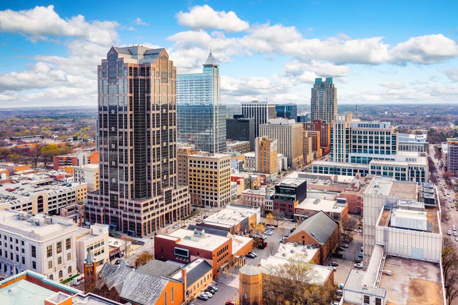 Aerial view of downtown Raleigh, North Carolina, featuring a mix of modern glass skyscrapers and historic brick buildings under a bright blue sky with scattered clouds.