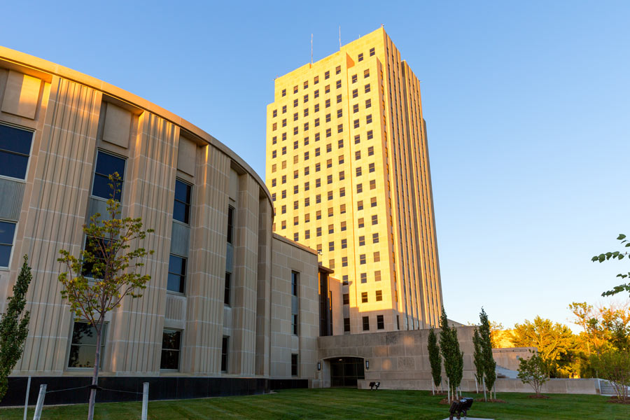"Modern metal-clad government building adjacent to tall Art Deco tower with golden exterior, small landscaped lawn with young trees in foreground, clear blue sky.