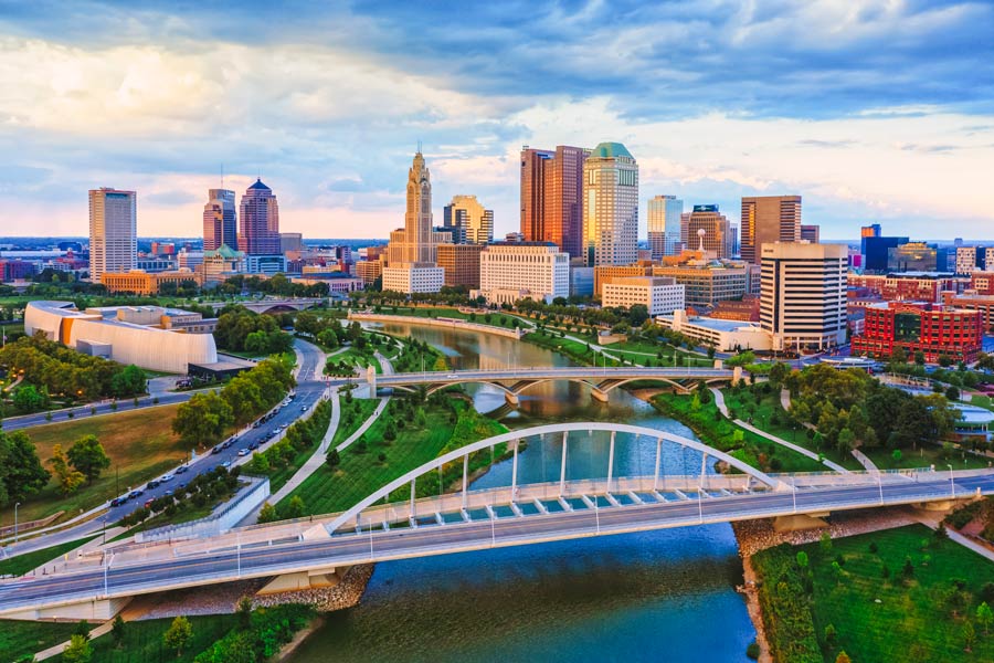 Columbus, Ohio skyline at golden hour along the Scioto River, with a white arch bridge in the foreground and the Scioto Mile riverfront park below downtown.