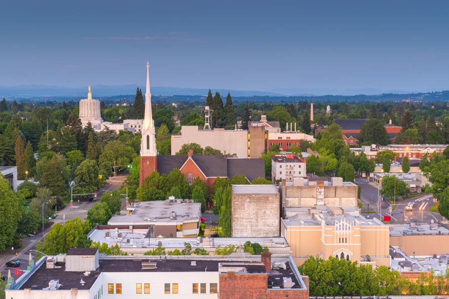 Aerial view of downtown Salem, Oregon at dusk, with the Oregon State Capitol's gold pioneer statue visible at left and a church steeple rising above the tree-lined city.