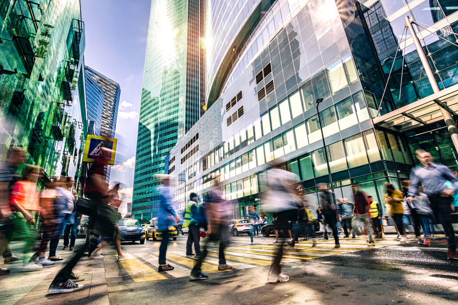 Blurred motion shot of a crowd of pedestrians crossing a busy urban intersection surrounded by tall glass skyscrapers in bright sunlight.