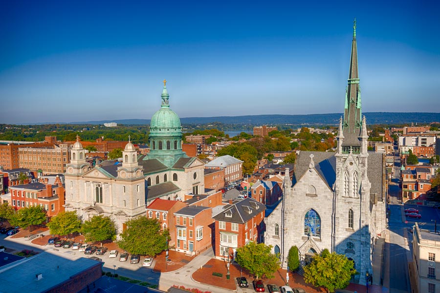 Aerial view of downtown Harrisburg, Pennsylvania featuring historic stone churches with a copper dome and tall steeple, with the Susquehanna River visible in the background.