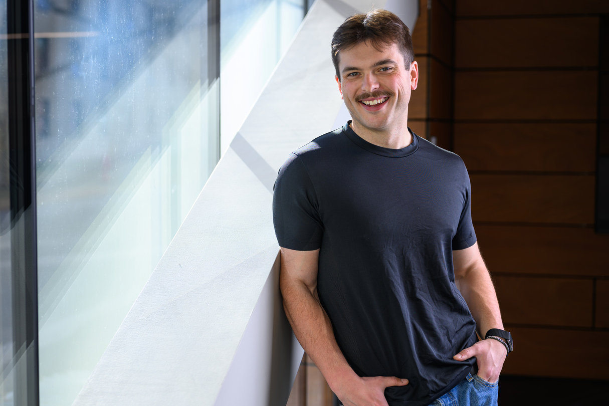 A man smiles while leaning against a wall near a large window.