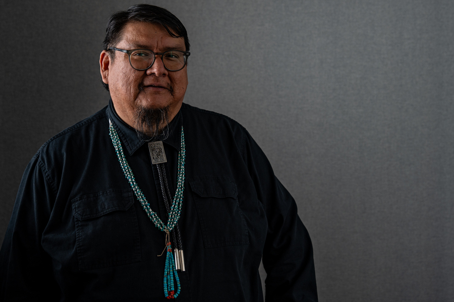Portrait of a man wearing glasses, a black shirt, and layered turquoise and coral bead necklaces with a silver bolo tie against a neutral gray background.
